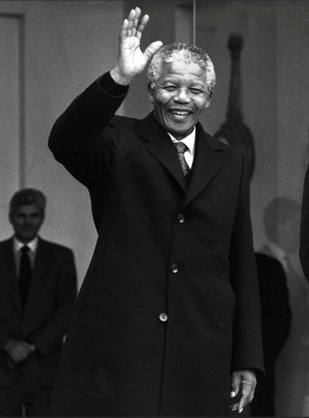 Black-and-white photograph of Nelson Mandela smiling and raising his hand in greeting